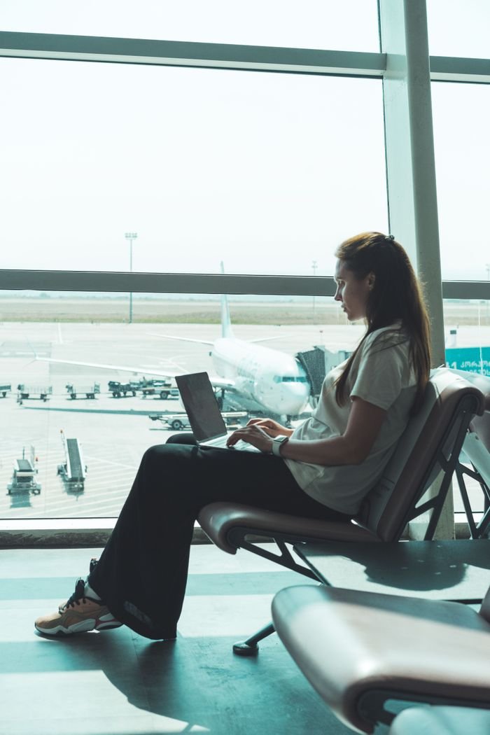 Side view of woman using laptop at airport, airplane visible outside through large glass windows.